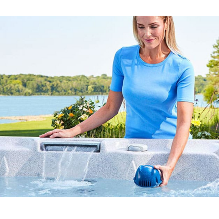 A woman in a blue shirt expertly uses the FROG @ease Swim Spa Sanitizing System in the hot tub by the lake, ensuring crystal-clear mineral water through advanced SmartChlor Technology.
