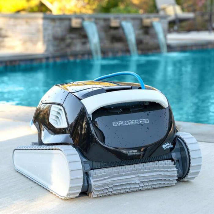 On a poolside patio with waterfalls and a shimmering pool in the background, the Maytronics Dolphin Explorer E30 with WiFi Robotic Pool Cleaner 99996241-XPI utilizes advanced Dolphin technology to deliver efficient pool cleaning. Effortlessly control it using the MyDolphin Plus app.