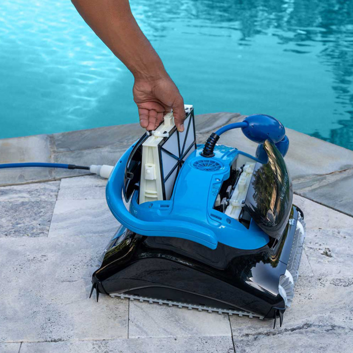Person replacing the filter of a blue and black Maytronics Dolphin Nautilus CC Plus Cleaner 99996406-PCI (with Wi-Fi) on poolside tiles near a swimming pool.
