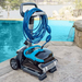 On the poolside patio, a Maytronics Dolphin Nautilus CC Plus Cleaner 99996406-PCI (with Wi-Fi) glides quietly with a coiled blue hose beside it, all set against a serene lake view. For added convenience, control and monitor your pool robot effortlessly through the MyDolphin Plus app.