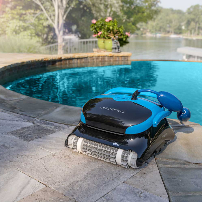 A Maytronics Dolphin Nautilus CC Plus Cleaner 99996406-PCI (with Wi-Fi) on a stone deck next to a clear blue swimming pool, with a lake and greenery in the background. Easily controlled via the MyDolphin Plus app, this pool robot ensures your oasis remains spotless.