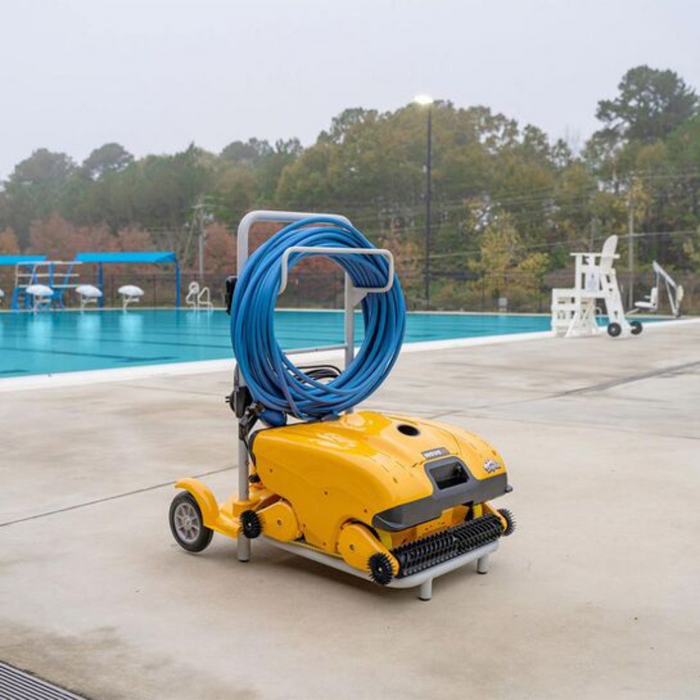 The Maytronics Dolphin Wave 140 Commercial Robotic Pool Cleaner 99997140-US, featuring a vibrant yellow design and equipped with a blue hose, rests on the poolside deck by an outdoor swimming pool. It provides heavy-duty scrubbing while being energy-efficient.