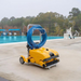 The Maytronics Dolphin Wave 140 Commercial Robotic Pool Cleaner 99997140-US, featuring a vibrant yellow design and equipped with a blue hose, rests on the poolside deck by an outdoor swimming pool. It provides heavy-duty scrubbing while being energy-efficient.