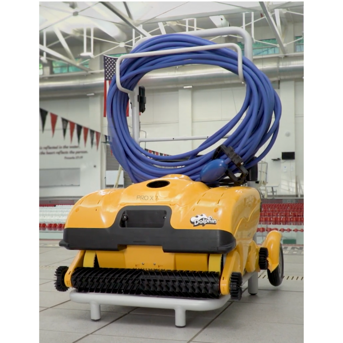 The Maytronics Dolphin Wave 140 Commercial Robotic Pool Cleaner 99997140-US, known for its heavy-duty scrubbing capability, is positioned on a tiled floor with its coiled blue hose beside an indoor swimming pool.