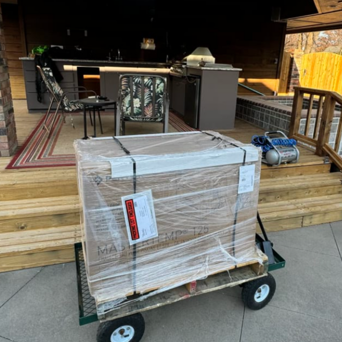 A large, wrapped package sits on a cart near an outdoor kitchen with black countertops and patterned chairs, possibly concealing cutting-edge innovations like the Pentair 125K BTU MasterTemp 125 Propane Heater with Cord EC-462025 461061 from Pentair Pool, which promises enhanced thermal efficiency.